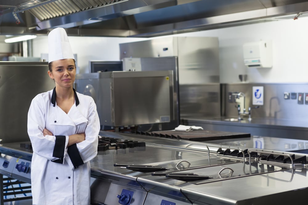 Young happy chef standing next to work surface arms crossed in professional kitchen